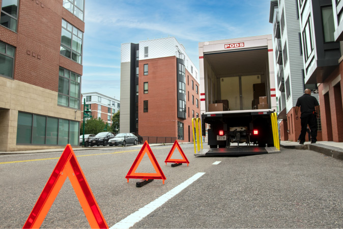 A PODS truck is parked outside a city building ready for loading and there are caution triangles placed in the street to warn drivers.