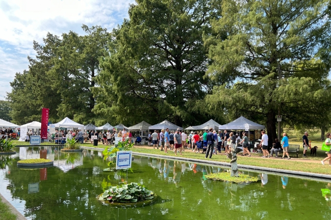 Residents living in St. Louis neighborhoods attend Art in the Park featuring a man-made pond at the center with pine trees in the background.