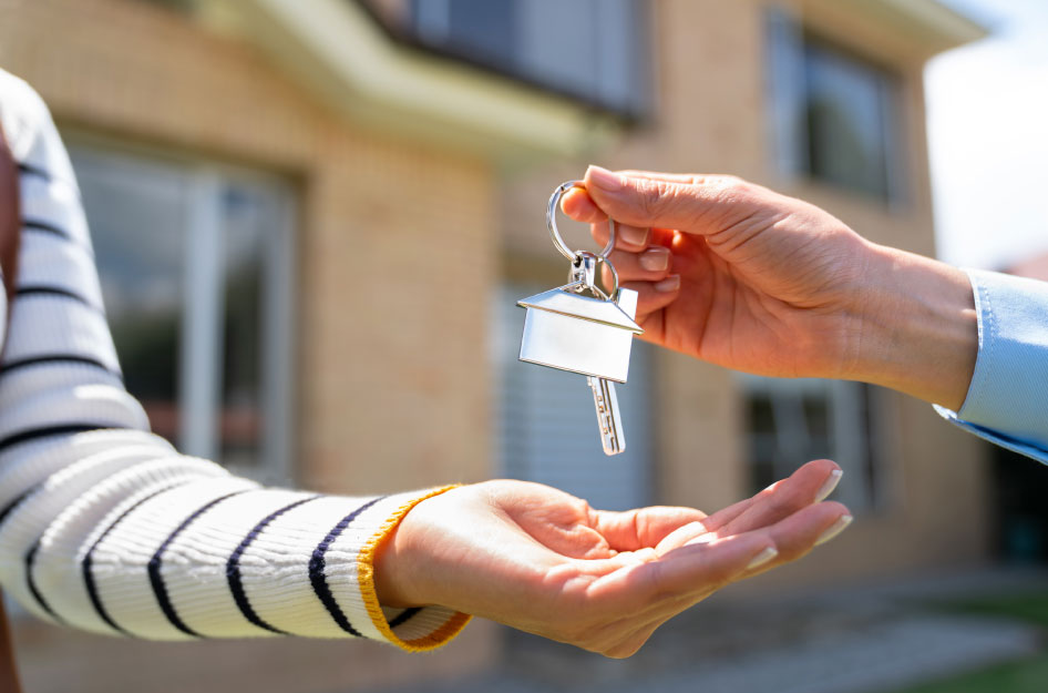Close-up of a realtor’s hand placing a set of house keys into a woman’s hand with her newly purchased house visible in the background.