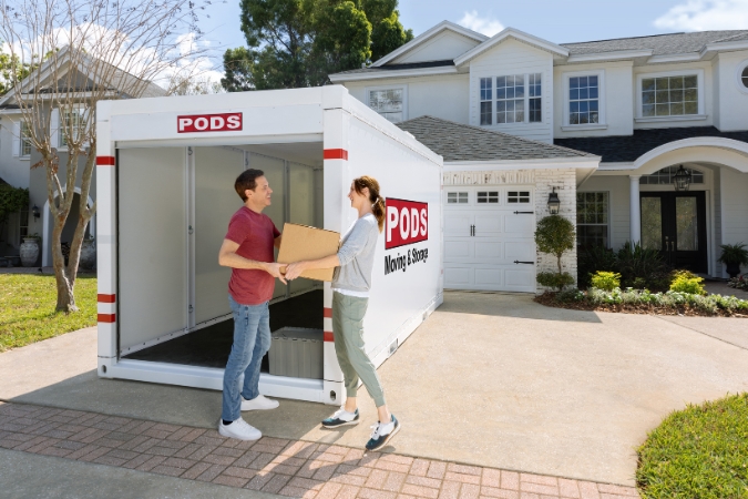 A couple moving during hurricane season hands each other a moving box as they unpack their PODS portable storage container in the driveway of their new home.