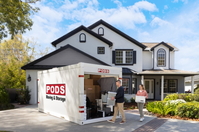 A couple is packing boxes and plastic bins into a PODS portable container in their driveway, while preparing to relocate to the best state to buy a house.