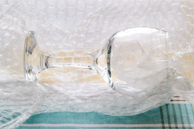 A wine glass is on its side atop a piece of bubble cushioning roll, as someone is preparing to wrap and pack it.
