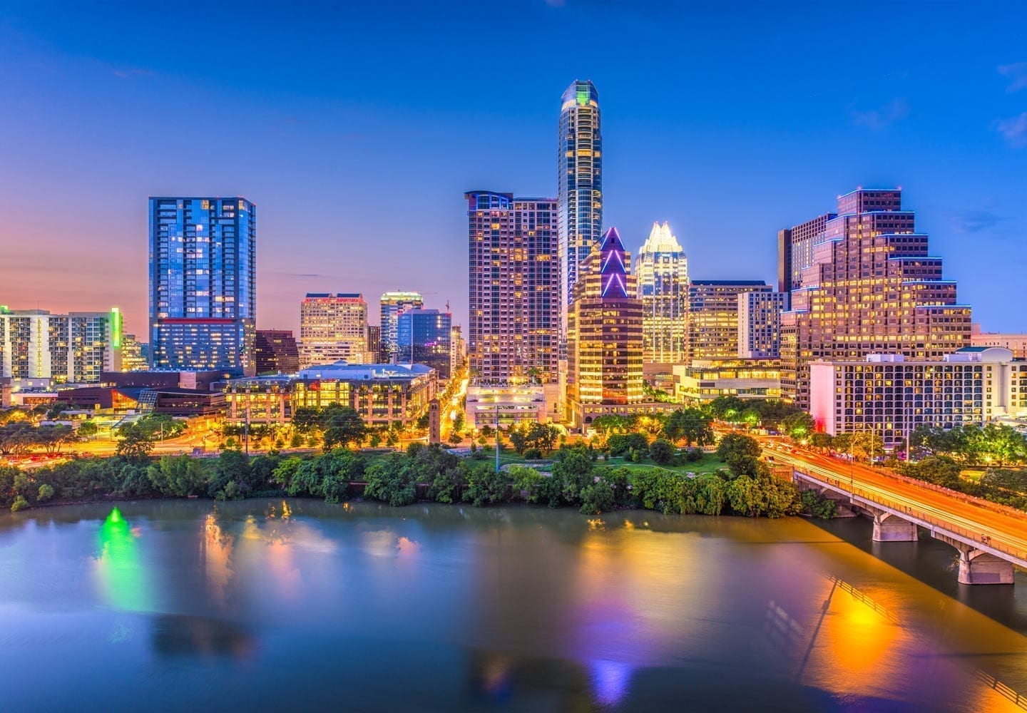 A night-time image of a city skyline at night. All the building are lit from the outside and it reflects off of the eater of a lake. 