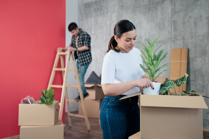 A woman boxes up live plants while a man stands on a ladder behind her, showing what not to put in a storage unit.
