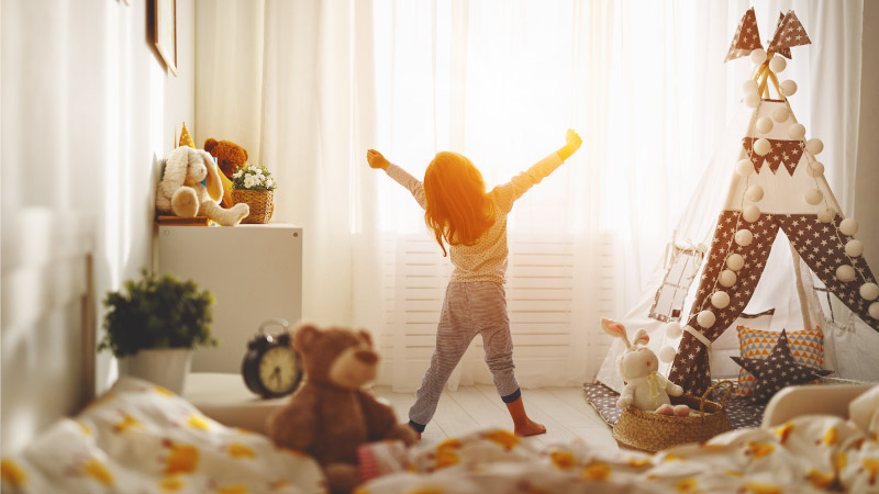 A little girl in her pajamas is stretching in front of her bedroom window as the morning sun pours into her room.