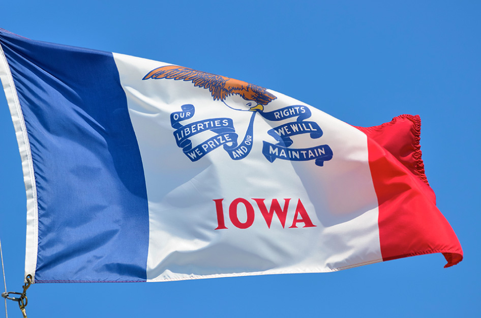 The Iowa state flag blowing in the wind in front of a clear blue sky.