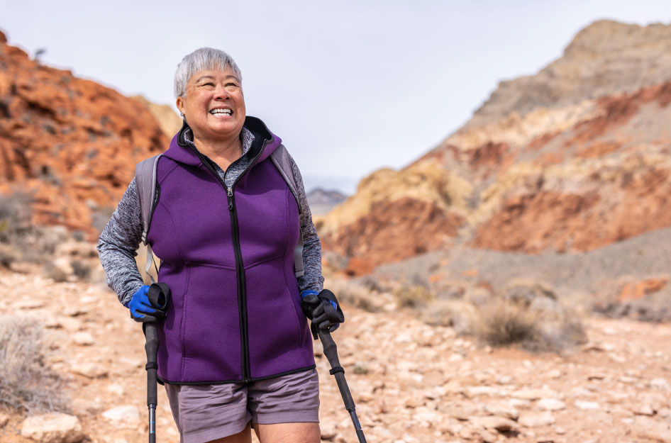A recently retired woman is wearing a purple fleece as she happily hikes through rocky terrain near her new Nevada home. She’s also wearing gloves and using hiking sticks.