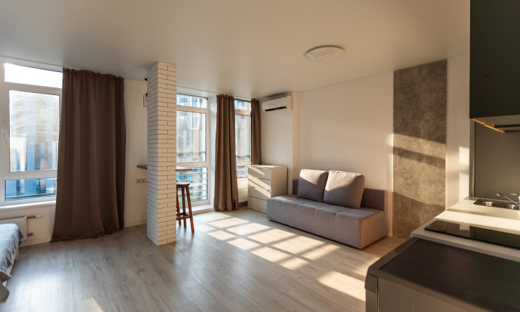 A studio apartment with only a few pieces of furniture. The space features tall skinny elements like windows, curtains, a brick column, and a vertical accent section of the wall. These design elements make the room seem larger. 