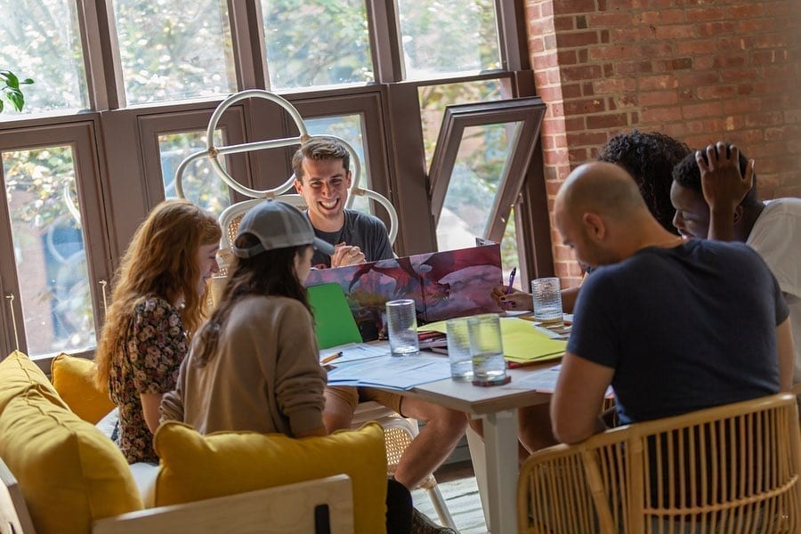 A group of friends gather in a loft apartment to celebrate moving. They are sitting around, laughing. There aare glasses of water on the table.