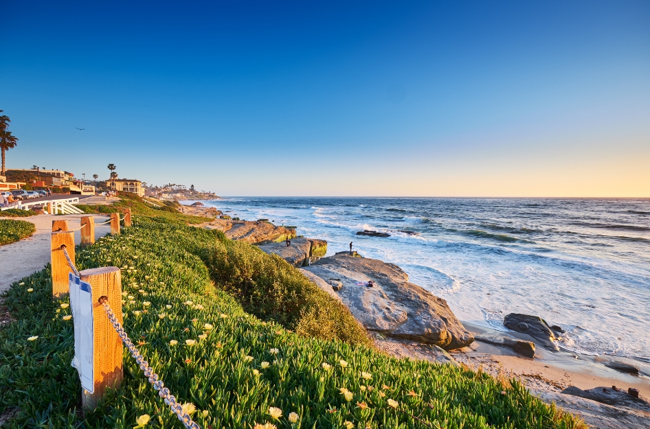 A sandy, fence-lined pathway with bright green grass leading down to a rocky shoreline, where waves crash against the beach in San Diego, California, one of the top states with the best weather.