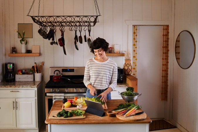  A young woman is chopping vegetables at her small kitchen island while she watches an instructional cooking video.