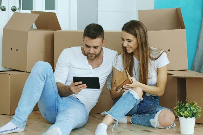 A young couple sits on the floor surrounded by moving boxes, smiling as they browse a tablet together.