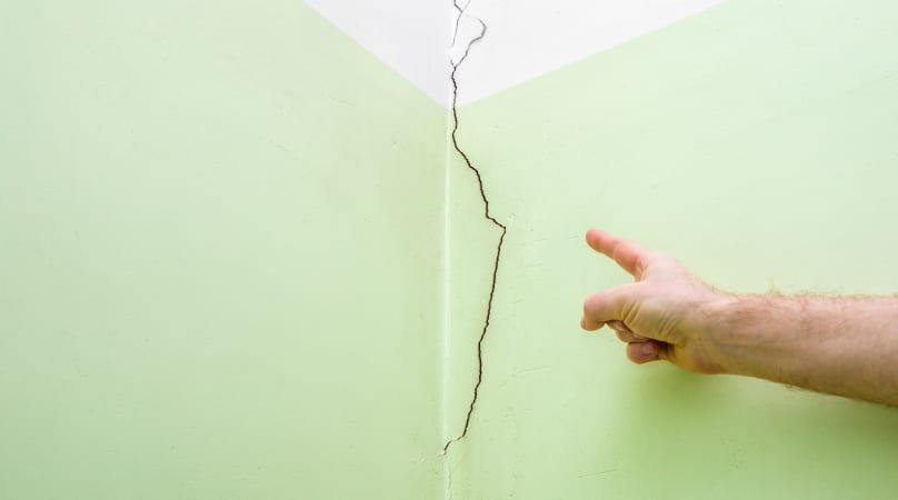 A crack runs down the middle of a green wall while a human finger points to the damage.