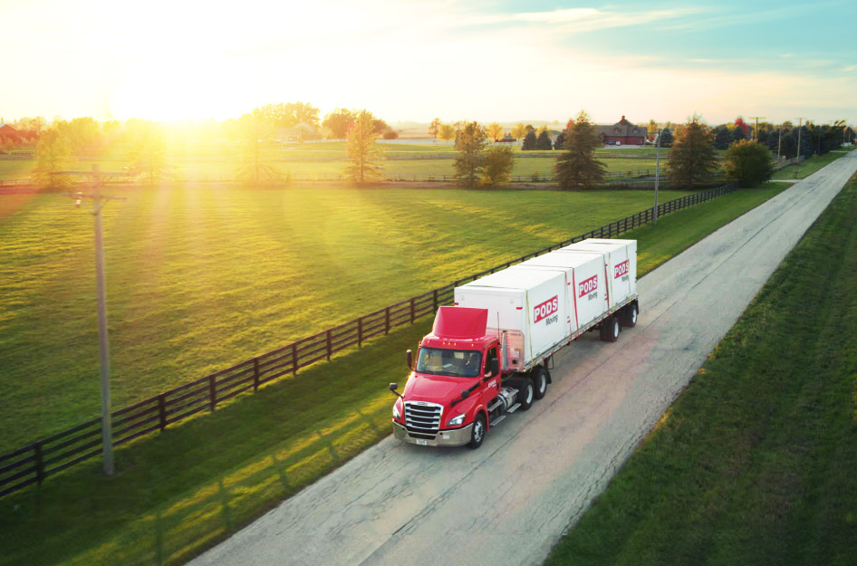 A PODS truck is transporting three PODS portable moving and storage containers along a country road past a green field as the sun sets.