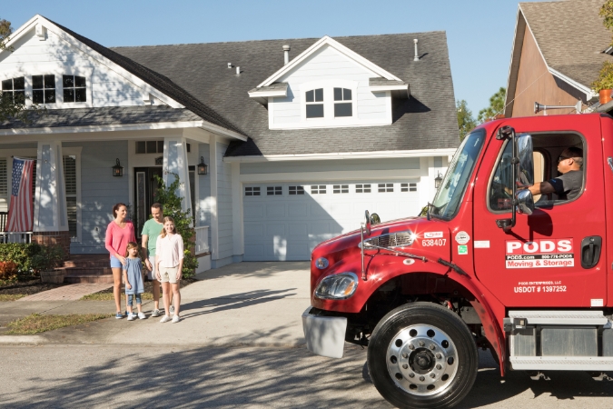 A family of four moving an estate stands outside their suburban home as a PODS delivery truck arrives with their PODS portable moving container.