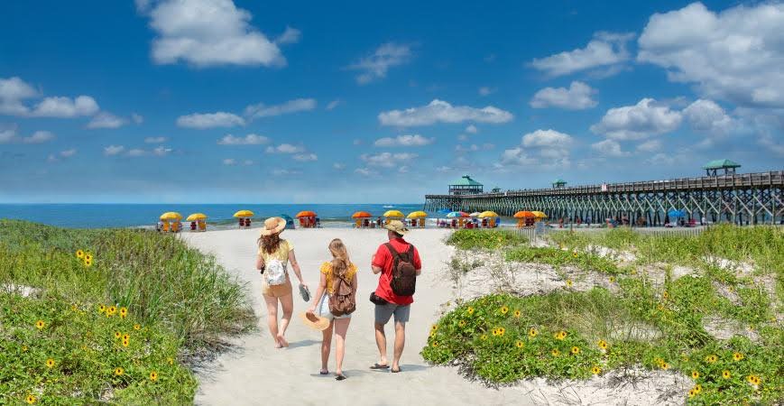 A family walks toward other beach-goers sitting under orange and yellow umbrellas near the pier at Folly Beach, South Carolina.