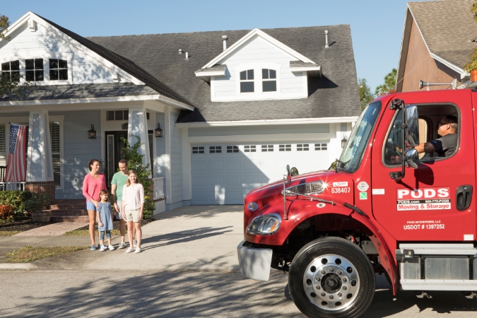 A family of four is standing in the driveway watching as a PODS truck arrives to deliver their portable moving container.