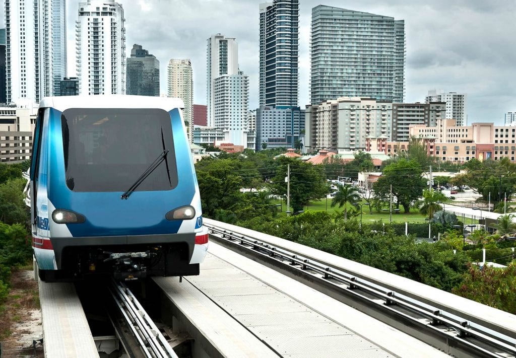 The cost of living in Miami includes the cost of transportation, but you can save money by using the Miami Metromover tram, seen here traveling on the tracks with the Miami city skyline visible in the background.