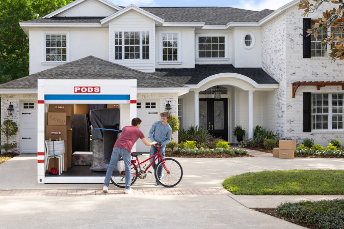 A teen is handing his bicycle to his father, who is packing a PODS container in the driveway of their home.