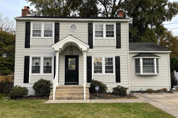 A colonial-style home with a one-level addition in Cherry Hill, New Jersey. The house features a small covered entryway, long black shutters, and a chimney on either end of the building.