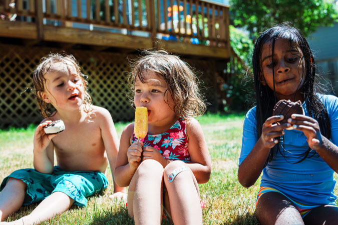 Three young kids are eating ice cream and popsicles on the lawn on a hot summer day.