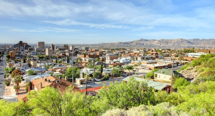 A treetop view of El Paso, Texas, on a summer afternoon, featuring residential buildings in the foreground, followed by taller city buildings, and a mountain range in the distance. 