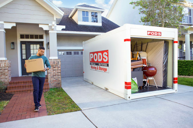 A man is carrying a moving box to a PODS container in his driveway, which is already nearly filled with household items to be put in storage.