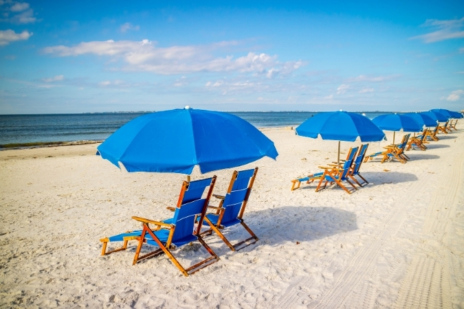 Blue beach lounge chairs and matching umbrellas line a picturesque beach in Fort Myers — just one of the many perks of living in Fort Myers, Florida.