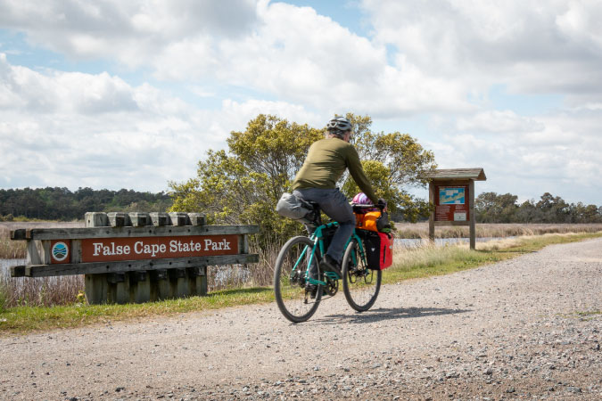 A man is biking on a trail in Virginia Beach’s False Cape State Park with his small child sitting in a seat secured to the front of his bike