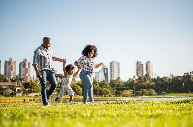 A father, mother, and daughter walk hand-in-hand through a park in a city.