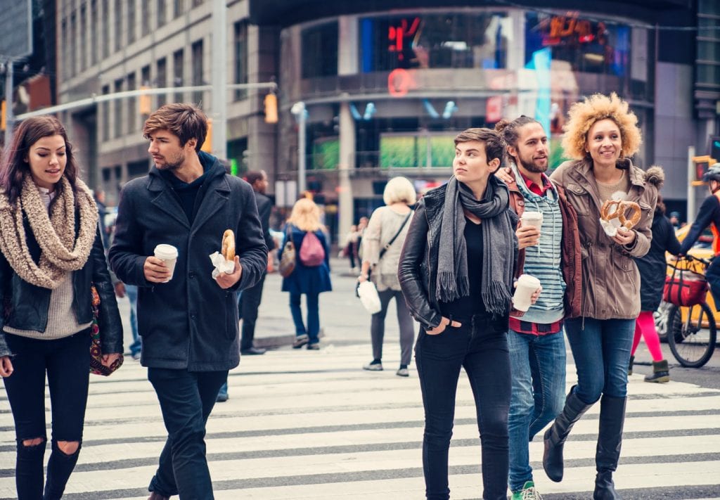 A busy crosswalk in Manhattan. There are lots of people walking in each direction — some carrying food, some carrying bags, some carrying on a conversation. Typical NY buildings can be seen in the background.