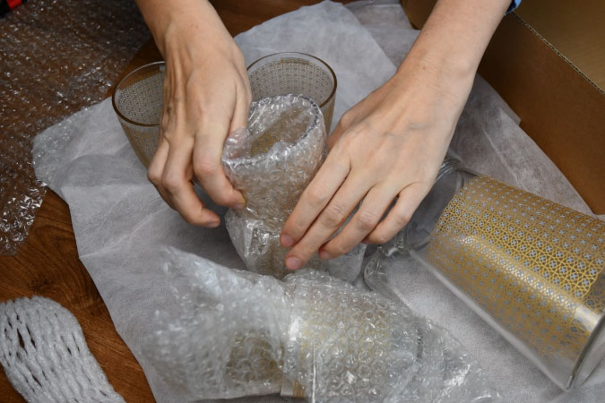 Close-up of a pair of hands using bubble cushioning roll to wrap up a drinking glass. There’s another glass that’s already been wrapped up, and two others and a pitcher that still need to be wrapped.