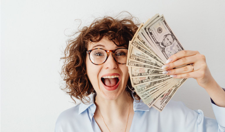 A young woman smiles a cheesy grin as she fans a handful of bills in front of her face. 