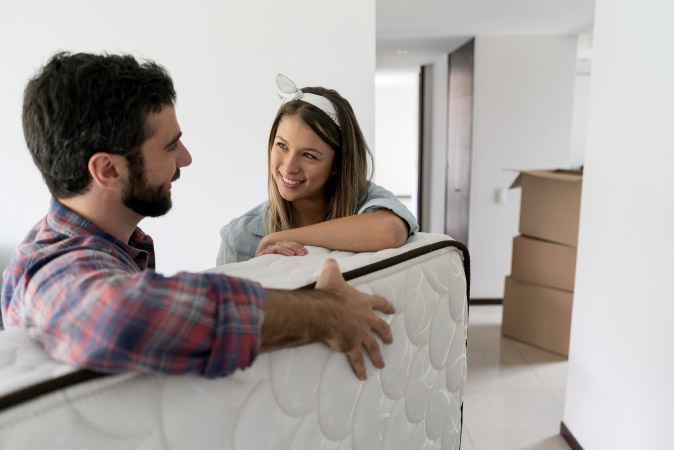 A couple pauses while moving a mattress, smiling at each other.
