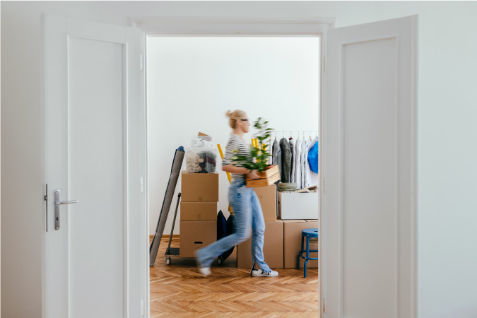A woman is walking quickly through her apartment as she packs for her upcoming move. There is a large pile of neatly stacked moving boxes and other personal items in the middle of her apartment.