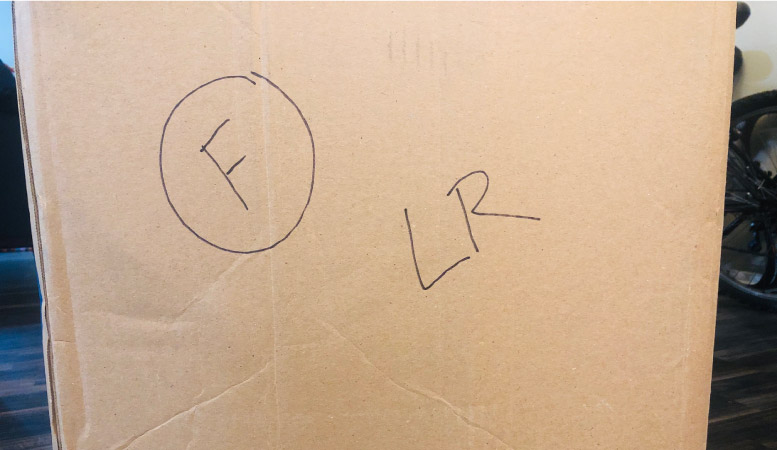 The side of a moving box with shorthand notes to indicate the contents are fragile (a circled “F”) and that the box will be unpacked in the living room (“LR”).