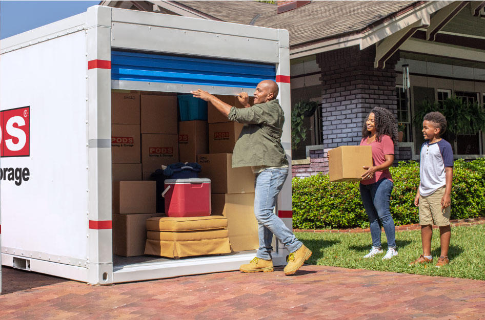 A soldier in civilian clothes is opening the door to a PODS portable moving container that he’s using for his DITY move. His wife is holding a moving box and his son is standing nearby.