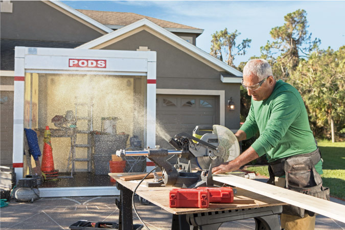 A contractor uses a miter saw to cut a wood board during an attic renovation project. There’s a PODS portable storage container in the driveway behind him filled with tools and materials for the project. 