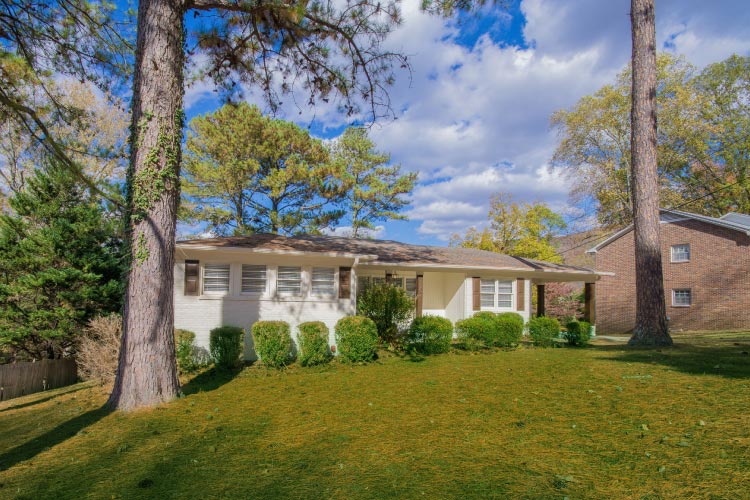  A beautiful white one-story home in Huntsville, Alabama, features a lush green lawn with a few tall pine trees. 