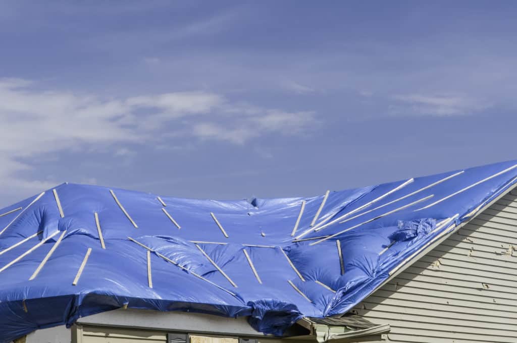 Blue plastic wrap, with wooden stays, stretched across roof of house damaged by a tornado
