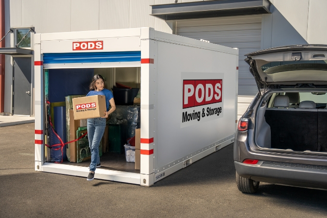 A woman is unloading a moving box from a PODS portable moving container that is positioned beside her vehicle, showcasing a moving cost that may qualify for a moving expenses tax deduction.