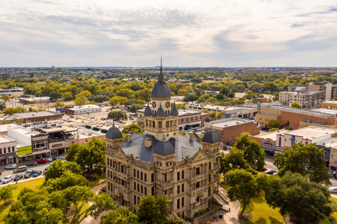 Aerial view of the square in Denton, Texas — one of the best cities near Dallas — featuring a spired city building and lush trees interspersed between other buildings and nearby neighborhoods.