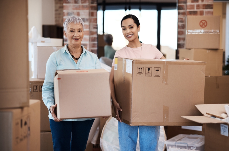 A woman is standing next to her mother, both holding moving boxes, as she navigates helping her elderly parents move into a new home.