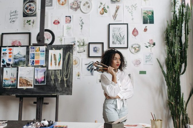 A young woman is standing with a handful of paint brushes beside a wall and easel displaying dozens of paintings and drawings.