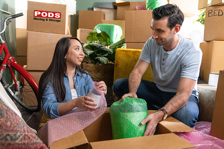 A young couple packs their PODS Boxes in order to prepare for their move to Dallas. One box is labeled “PODS Moving & Storage”. They are wrapping their delicate furniture items in pink and green bubble wrap. 