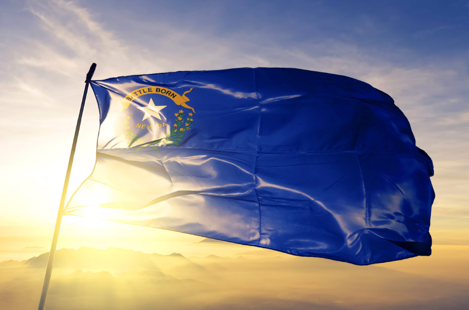 The state flag of Nevada flies high on a flag pole as the sun sets behind a mountain range in the distance.