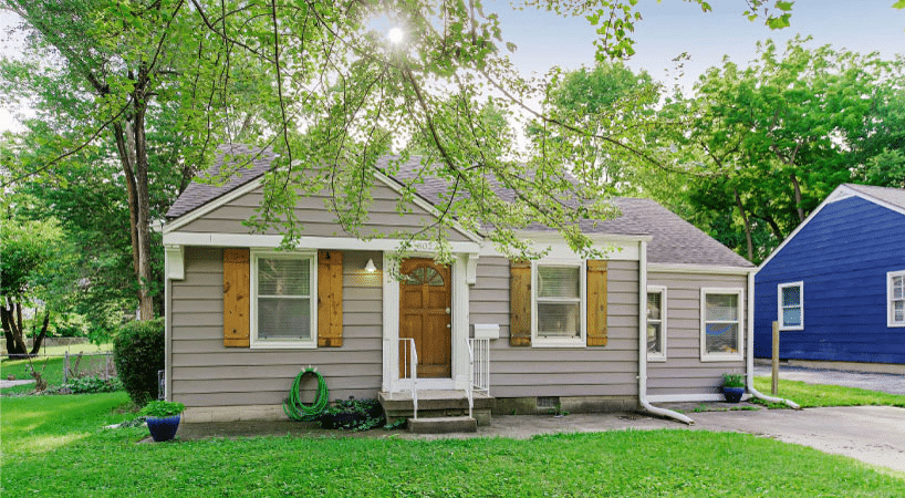 A small, single-family home in the Blue Hills neighborhood of Kansas City, Missouri. The exterior features gray siding with natural wood shutters. The sun is shining through the branches of a large tree in the front yard.