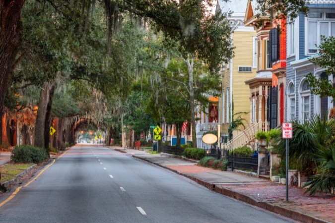A tree-lined residential street in Savannah, Georgia, featuring historic pastel homes and Spanish moss draping from oak trees across the way.