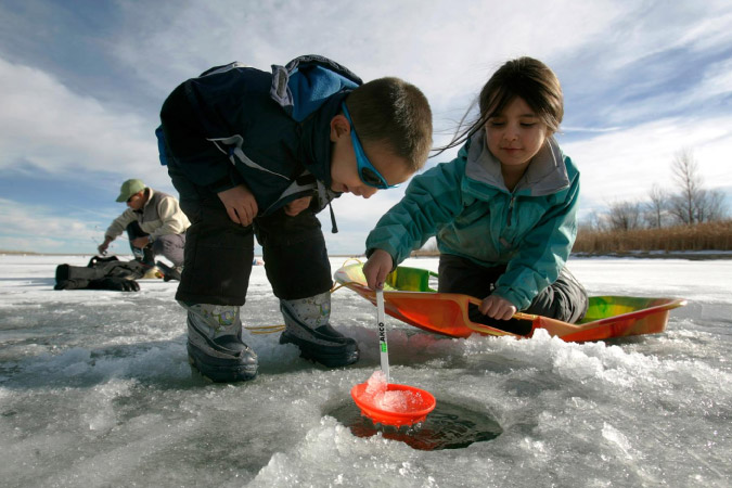 Two young kids living in Aurora, CO, use a plastic ladle to pull icy water out of a frozen pond while their dad prepares to ice fish behind them.