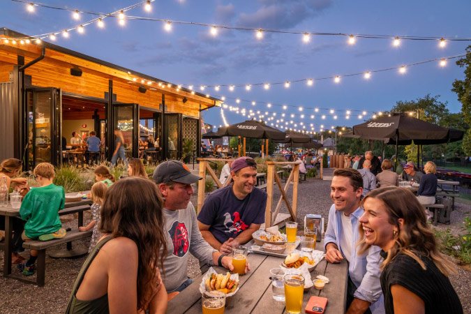Friends share drinks, food, and laughs at a picnic table in the garden of a St. Louis brewery.